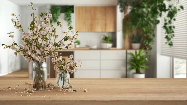 Wooden table, desk or shelf close up with branches of cherry blossoms in glass vase over blurred view of minimal kitchen and dining room, urban jungle interior design concept