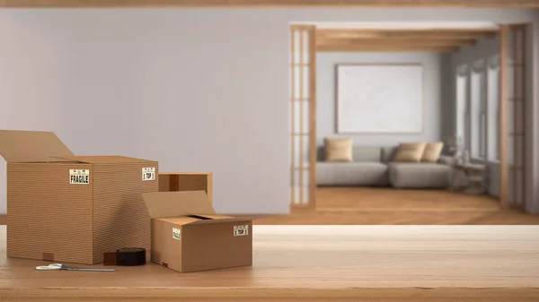 stock image Wooden table, desk or shelf with stack of cardboard boxes over blurred view of japandi living room in minimal style, interior design, moving house concept with copy space