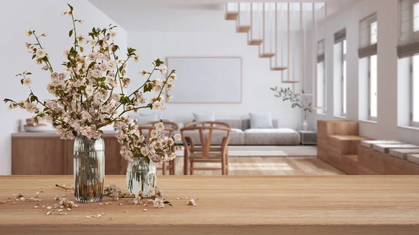 Wooden table, desk or shelf close up with branches of cherry blossoms in glass vase over minimalist dining and living room, minimal interior design concept