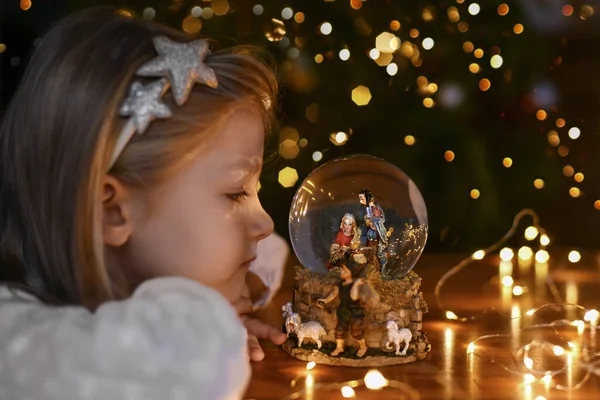 クリスマスツリーの近くにイエス キリストの誕生のシーンとガラスのボールを見て女の子 — ストック写真