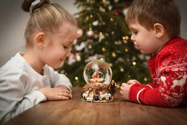 クリスマスツリーの近くにイエス キリストの誕生のシーンとガラスボールを見て子供たち — ストック写真