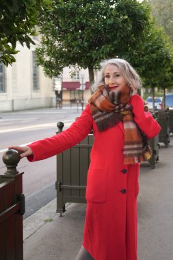 Kırmızılı, beyaz saçlı, kırmızı paltolu ve terracotta eşarplı bir kadın şehirde yürüyor.