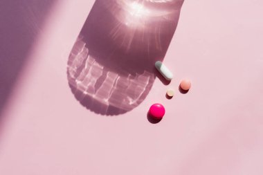 Tabletler ve haplar pembe arka plandaki bir bardak suyun gölgesinde yatıyor. Güçlü gölge ve yansıma efekti cam, üst görünüm, boşluğu kopyala