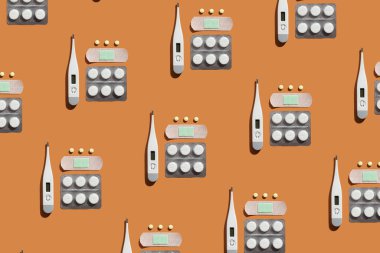 Turuncu arka planda tıbbi malzemelerin minimalist koleksiyonu. Beyaz dijital termometre, yeşil bloknotu olan yapışkan bandaj, yuvarlak sarı haplar ve beyaz haplar. Üst görünüm