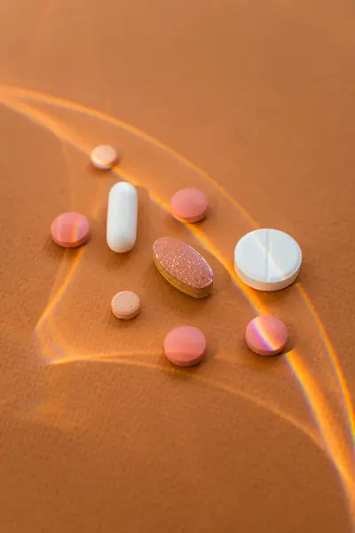 stock image Group of various pills or tablets with rainbow-like light reflections on brown background. White capsule, white round tablet, pink tablets, tablet with a glittery, small peach-colored round tablet