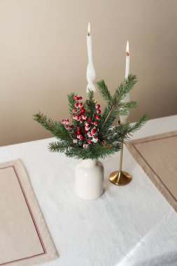 Christmas bouquet with red berries in a ceramic vase, two lit candles and two placemats on a table. The concept of preparation and table setting for the New Year's holiday. Hygge inspired home, cozy lifestyle clipart