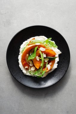 Krem peynirli ızgara balkabağı, ıspanak, roka ve badem gevreği beton arka planda siyah bir tabakta. Fırınlanmış Hokkaido balkabağı dilimleri, vejetaryen konfor yemekleri, en iyi manzara.