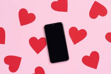 Boş ekranlı cep telefonunun üst görüntüsü ve pastel pembe arka planda kırmızı kağıt kalpler. Sevgililer Günü için maket şablonu. Aşk ve teknoloji kavramı, üst görünüm, kopyalama alanı