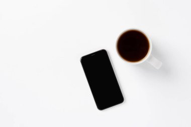 Beyaz fincanda beyaz akıllı telefon ve beyaz arka planda boş ekran var. Temizlik konsepti, tek renkli renk kombinasyonu. Metin veya tasarım için küçük arkaplan, üst görünüm