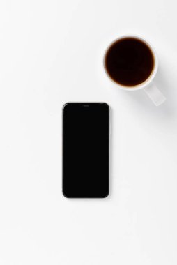 Beyaz fincanda beyaz akıllı telefon ve beyaz arka planda boş ekran var. Temizlik konsepti, tek renkli renk kombinasyonu. Metin veya tasarım için küçük arkaplan, üst görünüm