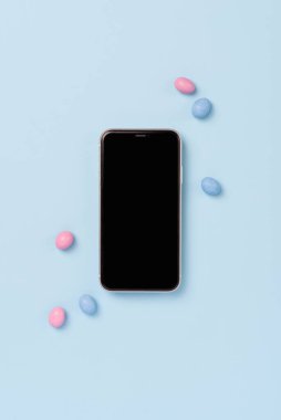 Siyah ekranlı cep telefonunun üst görüntüsü ve mavi arka planda mavi paskalya süprüntüsü. Paskalya için model şablonu. Yumurta şeklinde şekerlemeli akıllı telefon, yaratıcı paskalya konsepti, üst görünüm