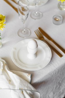 Mutlu Paskalyalar! Beyaz tabaklar, yumurta bardakları, altın çatal bıçaklar ve beyaz masa örtüsü üzerinde sarı çiçekler olan vazolar. Paskalya aile kahvaltısı ya da akşam yemeği konsepti
