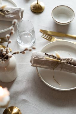 Bahar minimalist masası sıcak ve nötr renklerde. Paskalya bayramı masa dekorasyonu, beyaz tabaklar, altın çatal bıçak takımı, mumlar, yumurtalar ve vazodaki söğüt dalları. Paskalya aile yemeği ya da kahvaltı konsepti