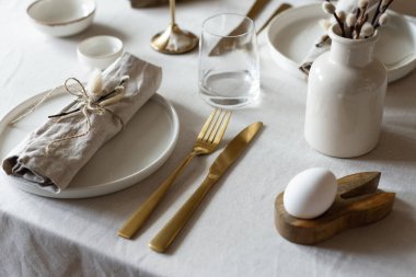 Bahar minimalist masası sıcak ve nötr renklerde. Paskalya bayramı masa dekorasyonu, beyaz tabaklar, altın çatal bıçak takımı, mumlar, yumurtalar ve vazodaki söğüt dalları. Paskalya aile yemeği ya da kahvaltı konsepti