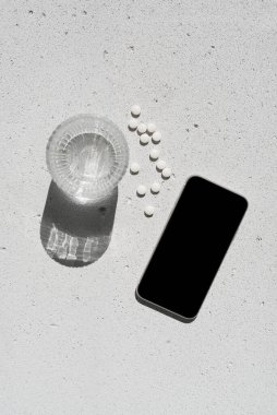 Boş ekranlı cep telefonu, beyaz vitamin hapları ve nötr beton arka planda saf suyla saf güneş ışığı gölgesi olan cam. Asgari sağlıklı yaşam tarzı konsepti. Saflık ve sadelik sahnesi, üst görünüm