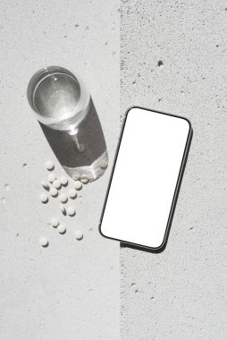 Boş ekranlı cep telefonu, beyaz vitamin hapları ve nötr beton arka planda saf suyla saf güneş ışığı gölgesi olan cam. Asgari sağlıklı yaşam tarzı konsepti. Saflık ve sadelik sahnesi, üst görünüm