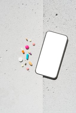 Boş ekranlı cep telefonu ve renkli vitamin hapları tarafsız beton arka planda, estetik güneş ışığı gölgesinde. Asgari sağlıklı yaşam tarzı konsepti. Saflık ve sadelik sahnesi, üst görünüm