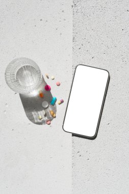 Boş ekranlı cep telefonu, renkli vitamin hapları ve nötr beton arka planda saf suyla saf güneş ışığı gölgesi olan cam. Asgari sağlıklı yaşam tarzı konsepti. Saflık ve sadelik sahnesi, üst görünüm