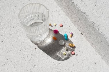 Renkli vitamin hapları ve nötr beton arka planda saf suyla birlikte cam ve estetik güneş ışığı gölgesi. Asgari güzellik, sağlık ya da eczane konsepti. Saflık ve sadelik sahnesi, üst görünüm
