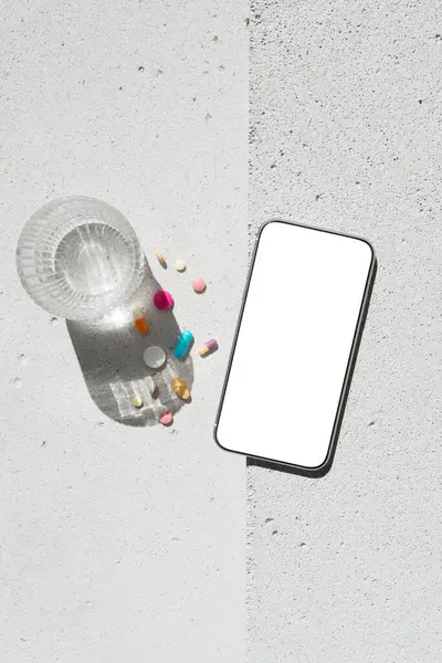 Boş ekranlı cep telefonu, renkli vitamin hapları ve nötr beton arka planda saf suyla saf güneş ışığı gölgesi olan cam. Asgari sağlıklı yaşam tarzı konsepti. Saflık ve sadelik sahnesi, üst görünüm