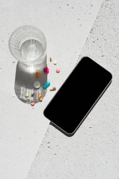 Boş ekranlı cep telefonu, renkli vitamin hapları ve nötr beton arka planda saf suyla saf güneş ışığı gölgesi olan cam. Asgari sağlıklı yaşam tarzı konsepti. Saflık ve sadelik sahnesi, üst görünüm