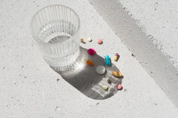 Renkli vitamin hapları ve nötr beton arka planda saf suyla birlikte cam ve estetik güneş ışığı gölgesi. Asgari güzellik, sağlık ya da eczane konsepti. Saflık ve sadelik sahnesi, üst görünüm