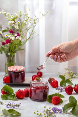 Hafif arka planda cam kavanozda taze ev yapımı çilek reçeli. Yanında birkaç taze çilek, yeşil çilek yaprağı ve çiçekler var..
