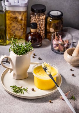Melted butter in jar surrounded by ingredients for cooking and herbs on white plate and table on light gray background.