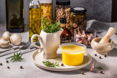 Melted butter in jar surrounded by ingredients for cooking and herbs on white plate and table on light gray background.