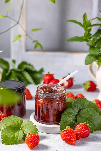 Hafif arka planda cam kavanozda taze ev yapımı çilek reçeli. Yanında birkaç taze çilek, yeşil çilek yaprağı ve çiçekler var..