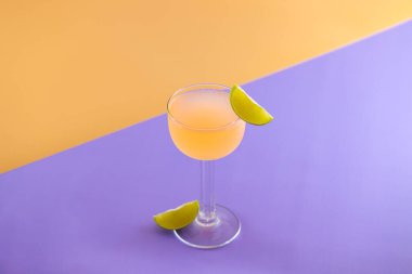 Pembe kokteyl, mor bir yüzeyde limon garnitürüyle duruyor.