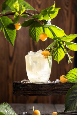 Glass filled with ice and a refreshing summer cocktail, garnished with yellow raspberries and rosemary, sits on a rustic wooden surface clipart