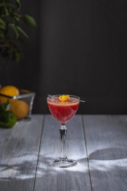 Kozmopolit karışımı limon bükülmesiyle süslenmiş bir coupe bardakta servis edilir, beyaz ahşap bir masada koyu bir arka planda.