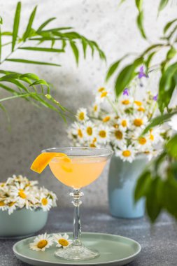 Portakal kokteyli, yeşil papatyalar ve yemyeşil çiçeklerle çevrili portakal kabuğu ile süslenmiş bir masada oturur ve ferahlatıcı bir yaz manzarası yaratır.