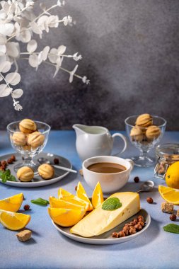Peynirli gri tabak, portakal dilimleri, fındık ve nane açık mavi bir masa örtüsünün üzerinde oturur, yanında da bir fincan kahve, kurabiye ve süt sürahisi, rahat bir kahvaltı ortamı yaratır.