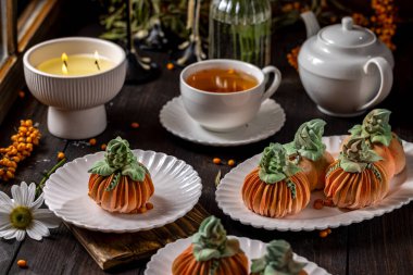 Balkabağı şekilli tatlılar, bir fincan çay ve yanan bir mumla birlikte beyaz tabaklarda servis edilir. Sıcak ve davetkar bir sonbahar atmosferini çağrıştırır.