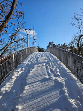 Köprüdeki karla kaplı patikayı kaplayan ayak izleri açık mavi gökyüzü altındaki iletişim kulesine doğru gidiyor.