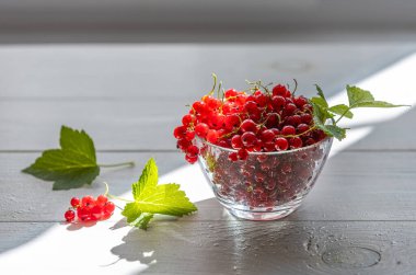 Olgun kırmızı üzümler cam bir kaseden dökülüyor, canlı yeşil yapraklarla birlikte, güneşli ahşap bir masada hoş bir yaz sahnesi yaratıyor.