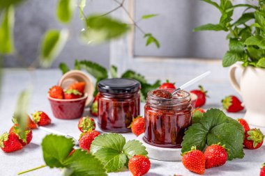İki kavanoz çilek reçeli. Birinin içinde kaşık var. Taze çilekler, yapraklar ve nane bitkisiyle çevrili.