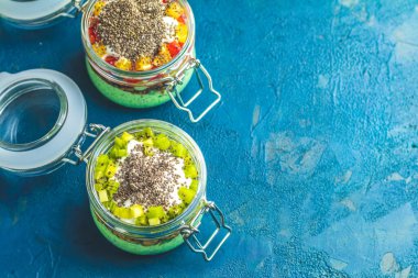 Yoğurt katmanlı, taze kivi ve mangoyla kaplı canlı yeşil chia tohumu pudingi ile dolu iki cam kavanoz.