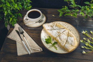 Bir tabak kahve, çatal bıçak ve peçeteyle servis edilen pudra şekerli elmalı turta.