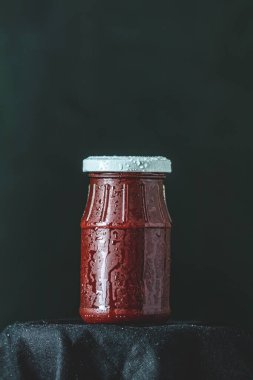 Yoğunlaşmayla kaplı kırmızı sos içeren küçük cam kavanoz siyah bir arkaplana karşı koyu bir bez üzerine oturur.