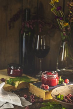 Cam kavanozda ev yapımı ahududu reçeli. Taze ahududu, nane yaprağı, bir şişe şarap, bir bardak şarap, eski kitaplar ve ahşap masadaki çiçekler.