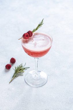 Canlı pembe kokteyl, taze ahududu ve biberiye dalıyla süslenmiş, şık bir coupe bardağında oturuyor, eğlenmeye hazır.