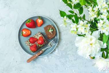 Taze çilekle süslenmiş çikolatalı kek, gri bir tabakta çatalla servis ediliyor, bir buket beyaz çiçeğin yanında.