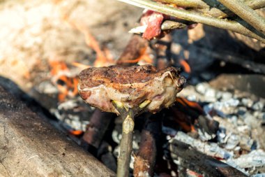 Kamp ateşinin üzerinde ahşap bir sopayla ızgara yapan bir et parçası kırsal alanda yemek pişirme deneyimi sunuyor.