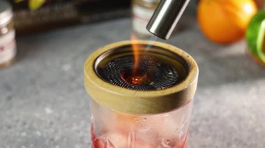 Barmen cam bir kavanozda kırmızı bir kokteyl içiyor meşaleyle ve ahşap kapakla kaplıyor.