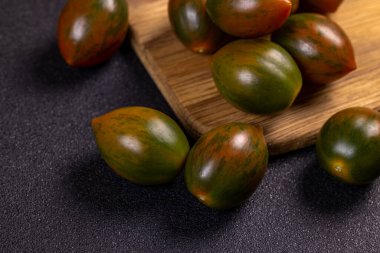 Olgun yeşil kumato domatesler koyu dokulu bir yüzeye ve ahşap kesme tahtasına saçılmış, görsel olarak çekici bir düzenleme yaratıyor.