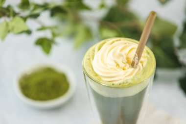 Krem şantili bir bardak matcha latte ve yeşil yapraklı bulanık bir arka planda toz yeşil çay.
