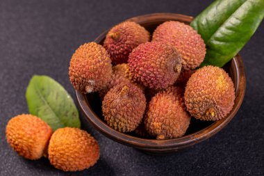 Olgun, dokulu deri ve yeşil yapraklı liçi, kırsal bir kasede sunuluyor ve koyu bir arkaplan karşısında canlı bir görüntü yaratıyor.